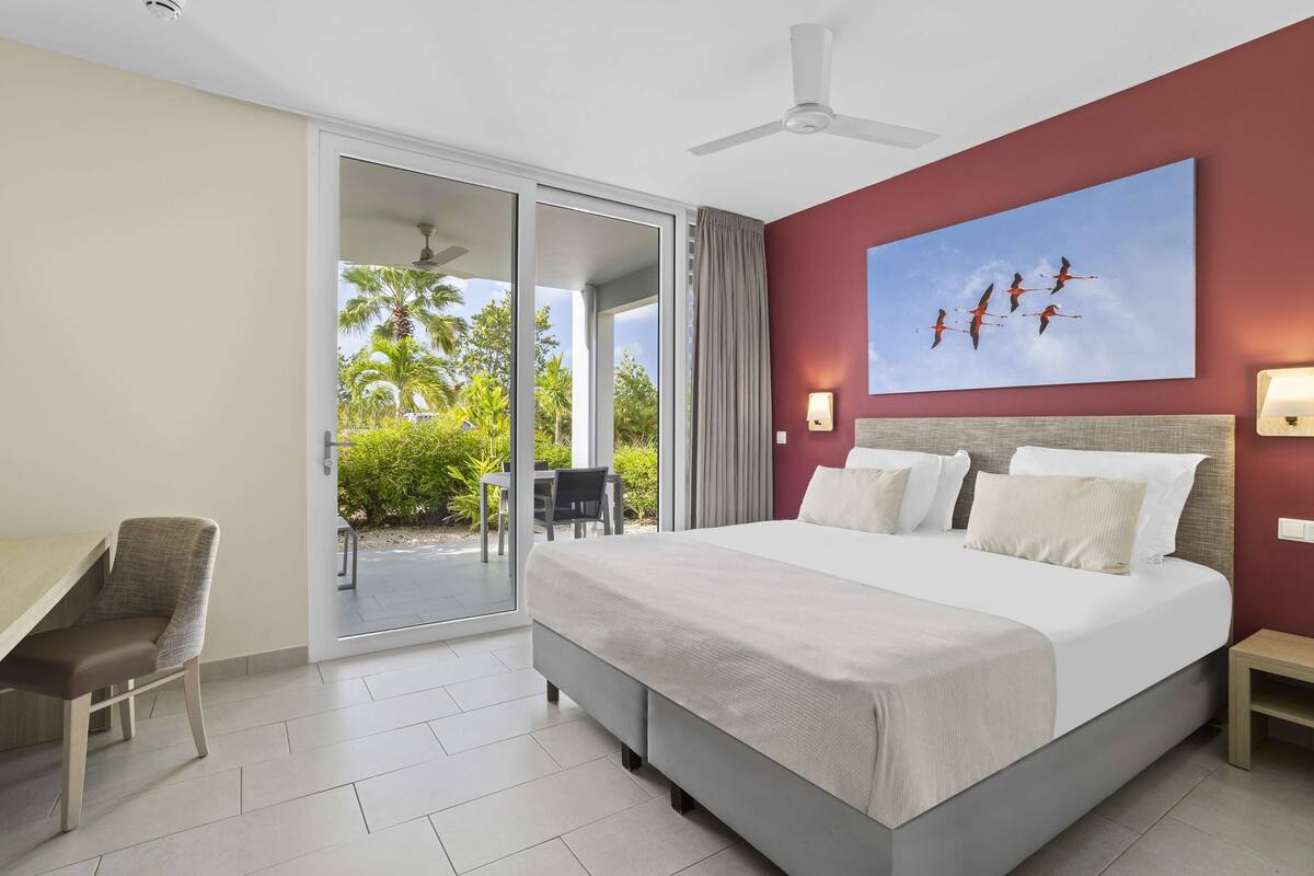 A hotel room with a bed, a desk, a chair, a ceiling fan, and a view of palm trees through the sliding glass door.