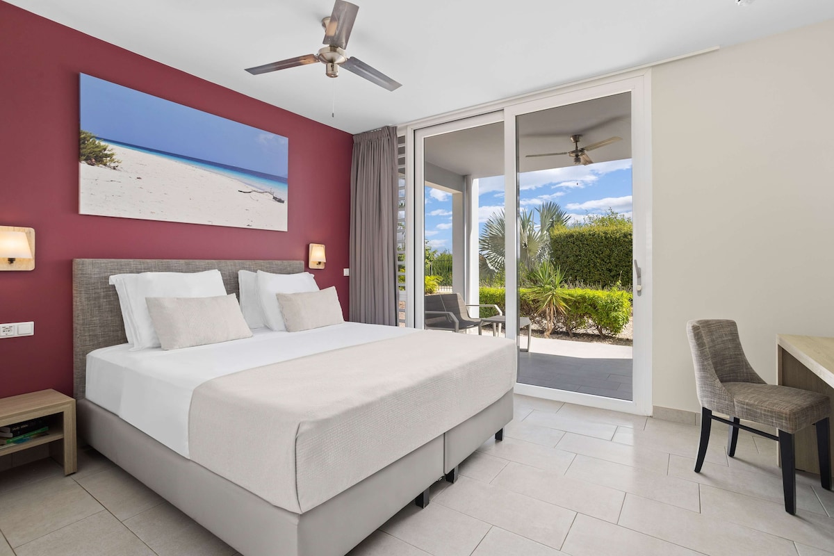 A bedroom with a large bed, a ceiling fan, a painting of a beach, and a sliding glass door leading to an outdoor area.