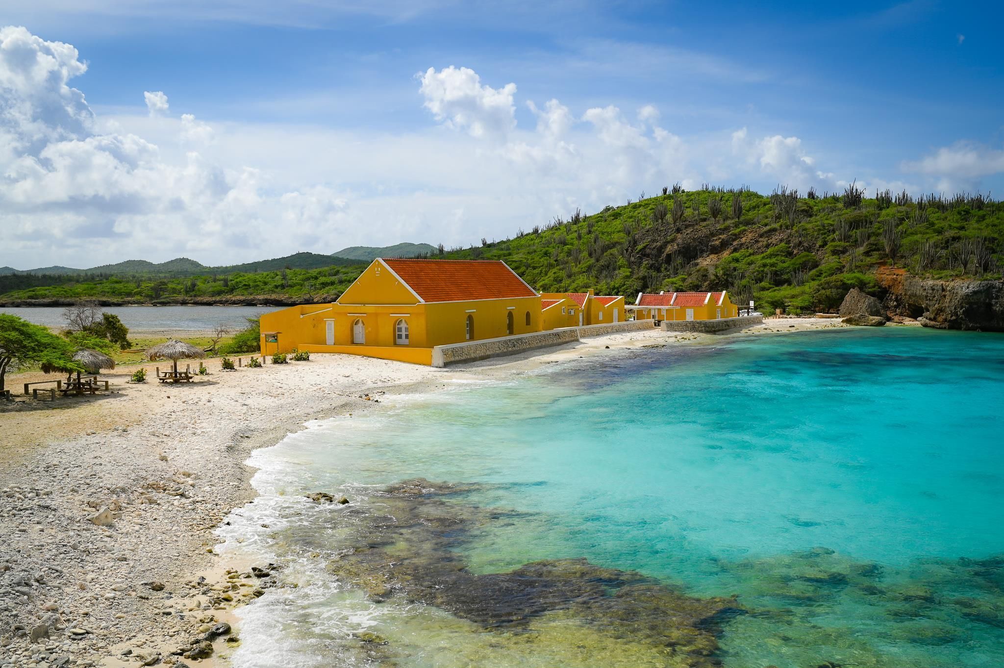 A coastal building with a red roof, situated on a sandy beach with clear turquoise waters.