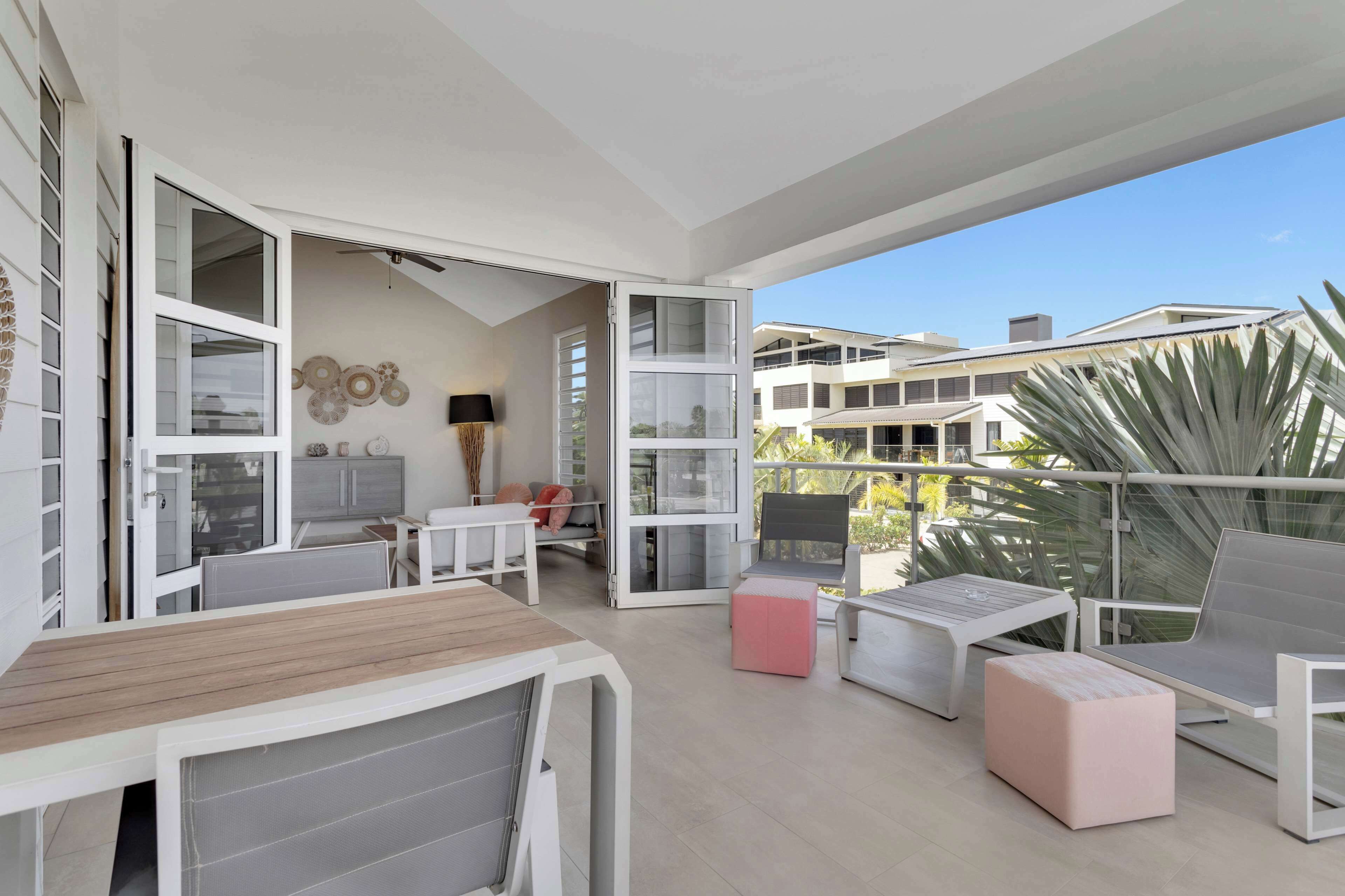 Un balcón moderno al aire libre con mobiliario blanco, una zona de comedor y vistas a edificios cercanos.