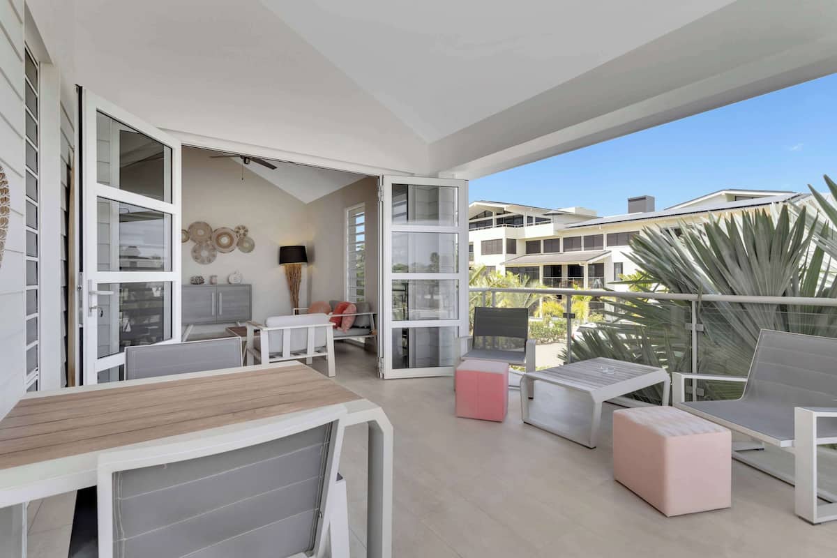 Un balcón moderno al aire libre con mobiliario blanco, una zona de comedor y vistas a edificios cercanos.