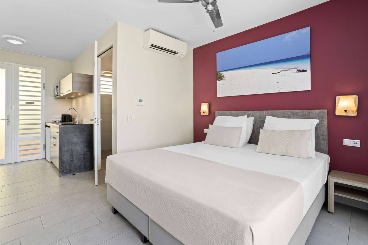 A bedroom with a bed, a painting of a beach, and a kitchenette.