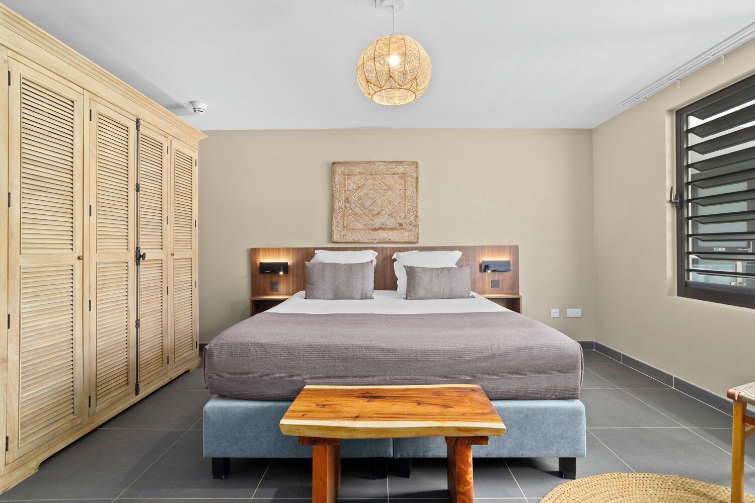 Une chambre à coucher comprenant un lit, des tables de chevet, une armoire, un luminaire suspendu et une fenêtre avec des stores.