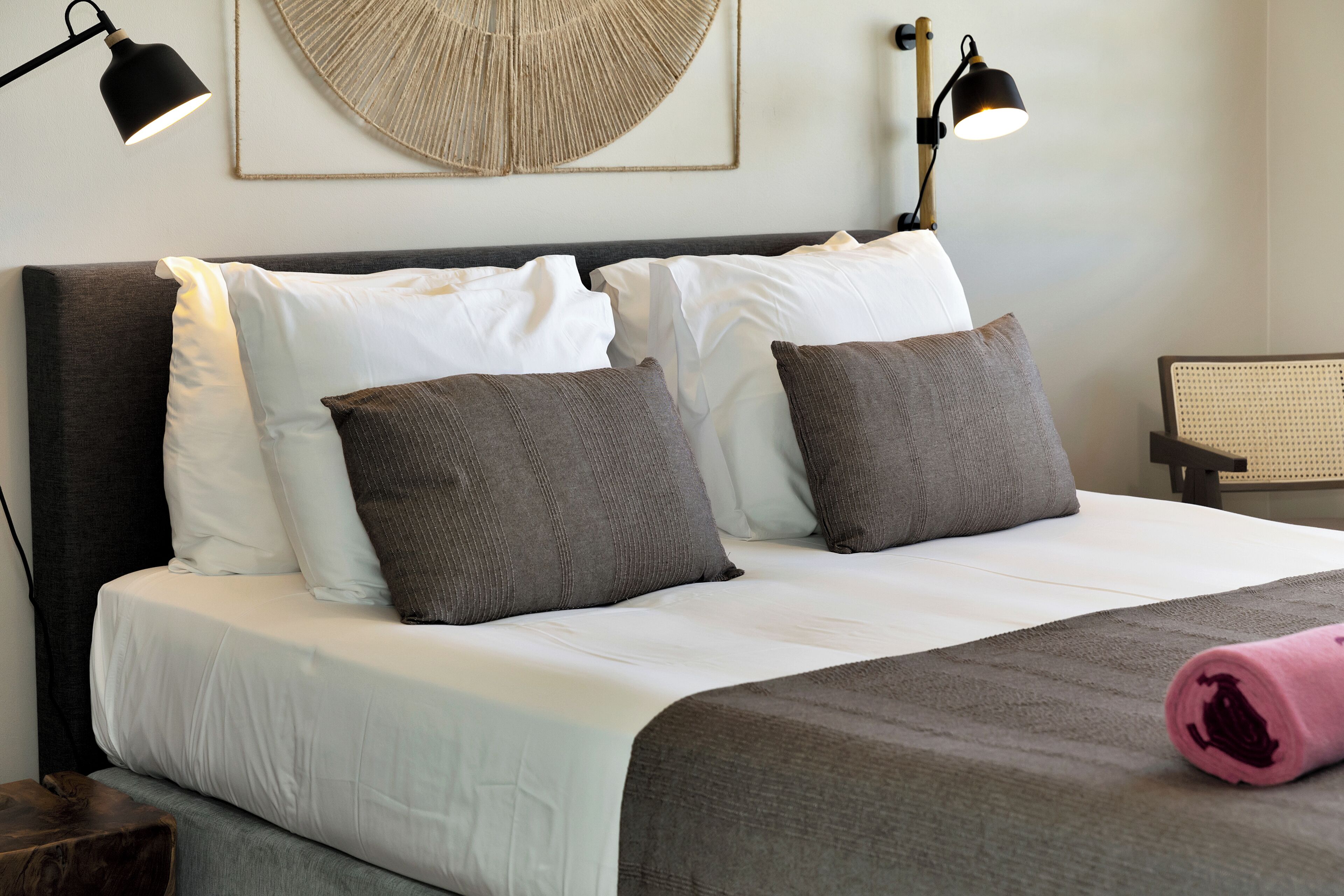 A neatly made bed with white and gray bedding, two dark gray pillows, and a pink towel. There are two wall-mounted lamps with black shades on either side of the bed.