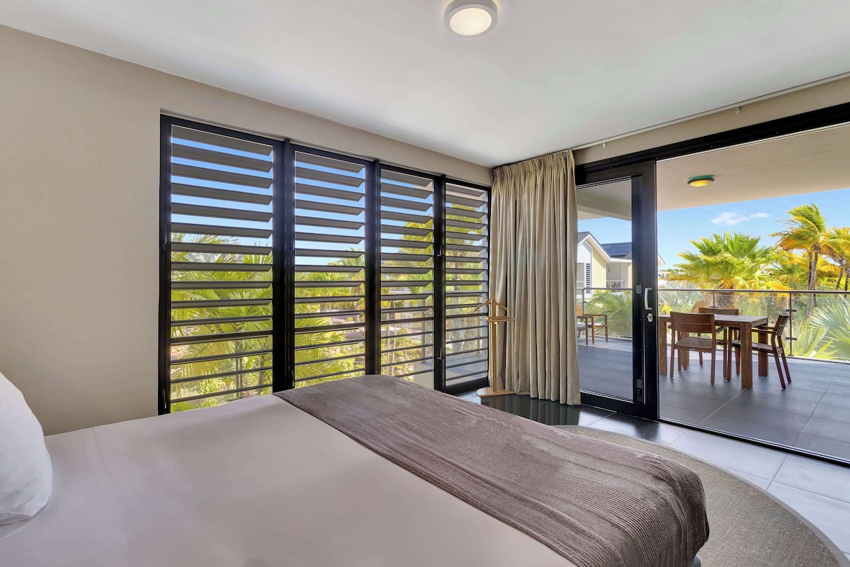 A hotel room with a large bed, a balcony with a table and chairs, and a view of palm trees.