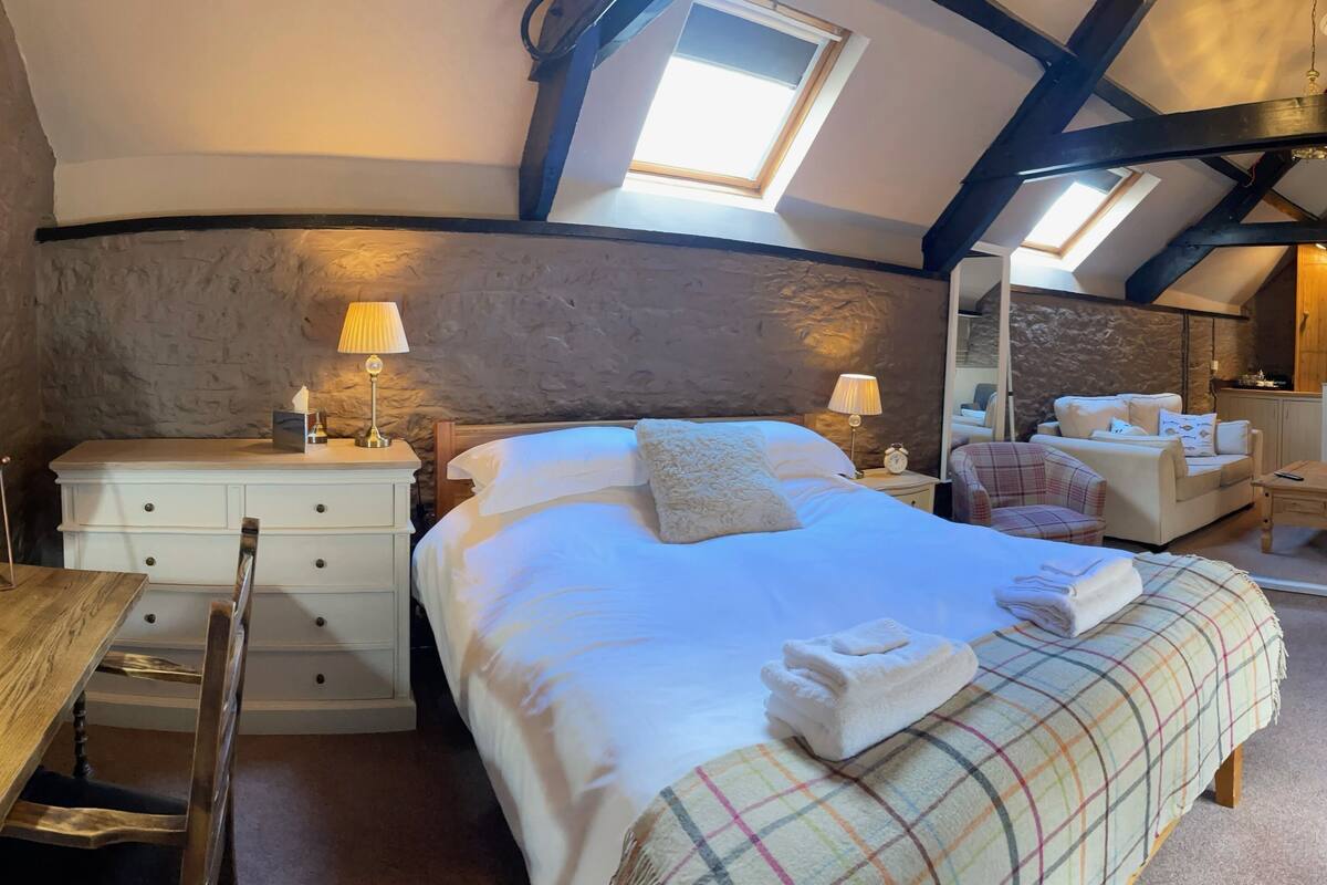 Ein gemütliches Schlafzimmer mit einem großen Bett, zwei Nachttischen mit Lampen, eine Holzkommode, einem kleinen Tisch und einem Stuhl.