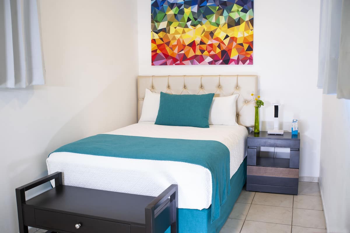 Une chambre d’hôtel bien rangée, avec un lit, une table de chevet et une œuvre d’art géométrique aux couleurs vives accrochée au mur.