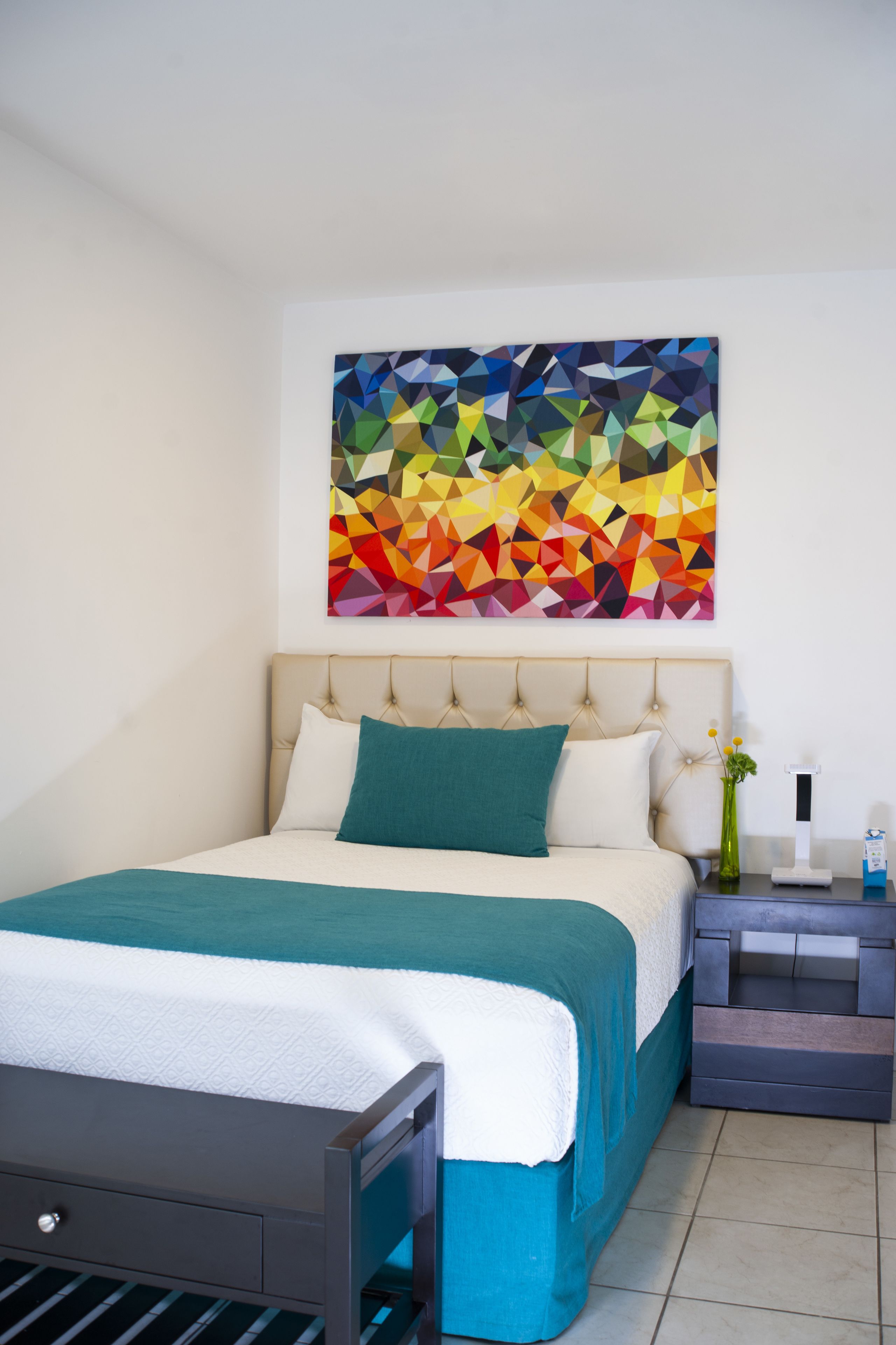 A bedroom with a bed, a nightstand, a vase with flowers, and a colorful geometric wall art.