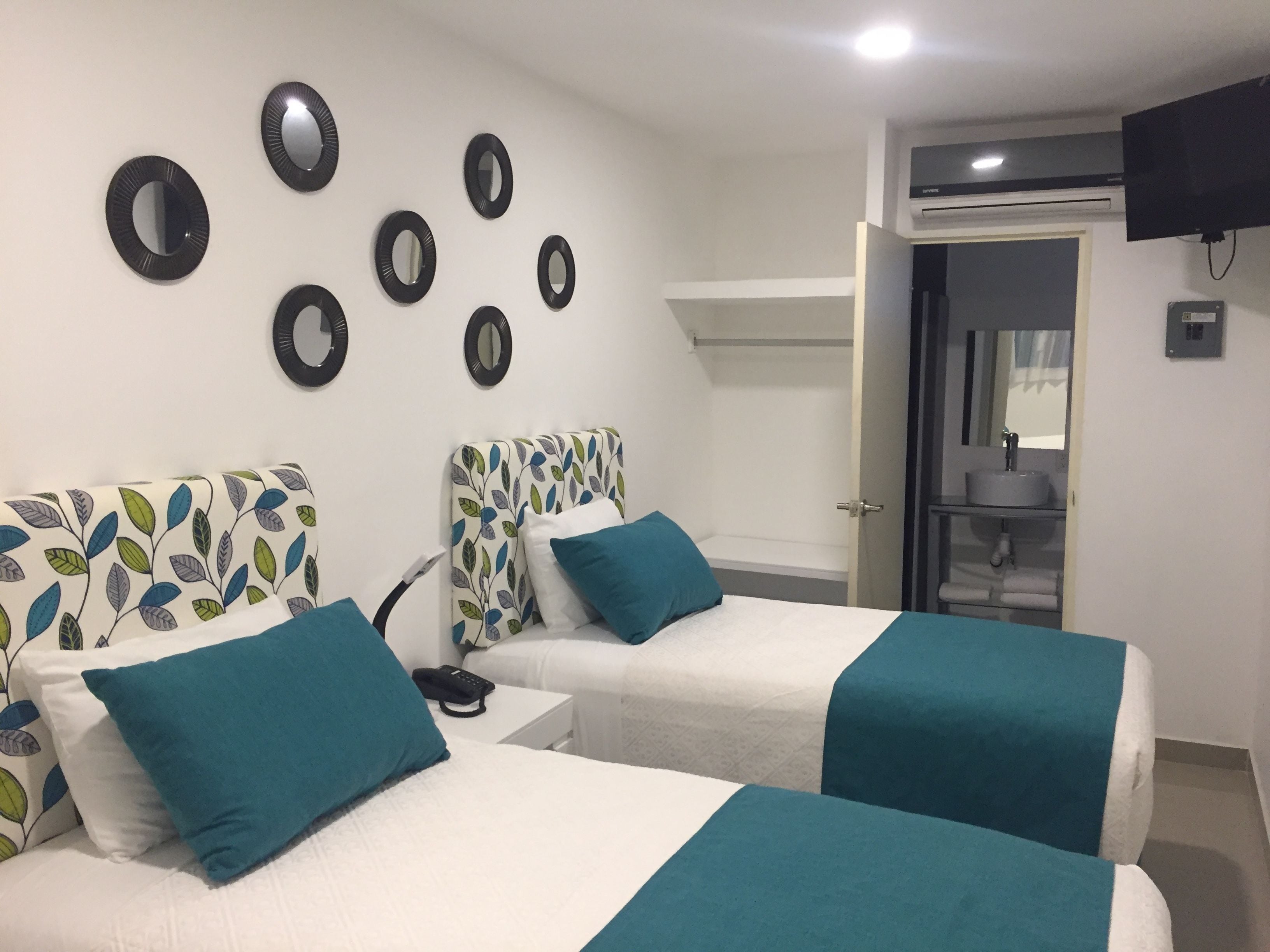 A hotel room with two beds, each with a teal bedspread and matching pillows. There are six circular mirrors on the wall, a television mounted on the wall, and a bathroom visible through an open door.