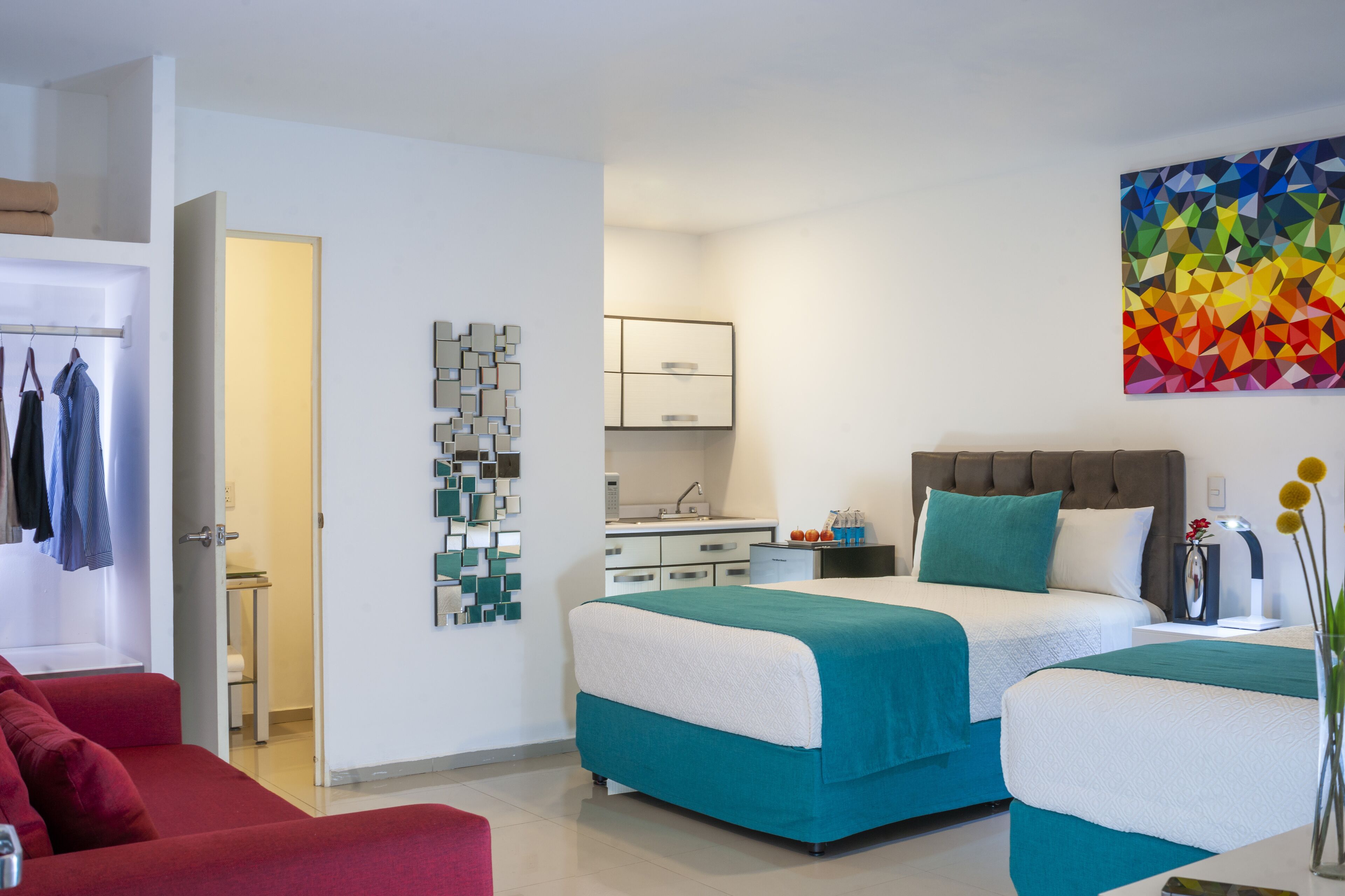 A modern hotel room with two beds, a red sofa, a kitchen area, and a colorful abstract painting.