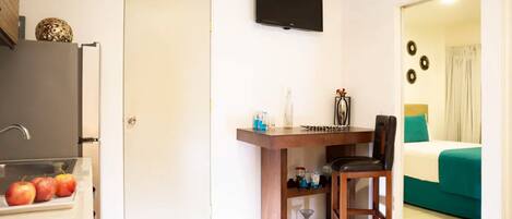 A hotel room with a bed, a TV mounted on the wall, a small table with a chair, and a refrigerator with apples on the counter.