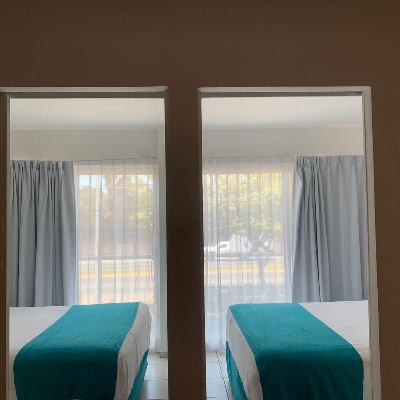 Una habitación con dos camas, cada una con colcha color turquesa, y ventanas amplias con cortinas translúcidas.
