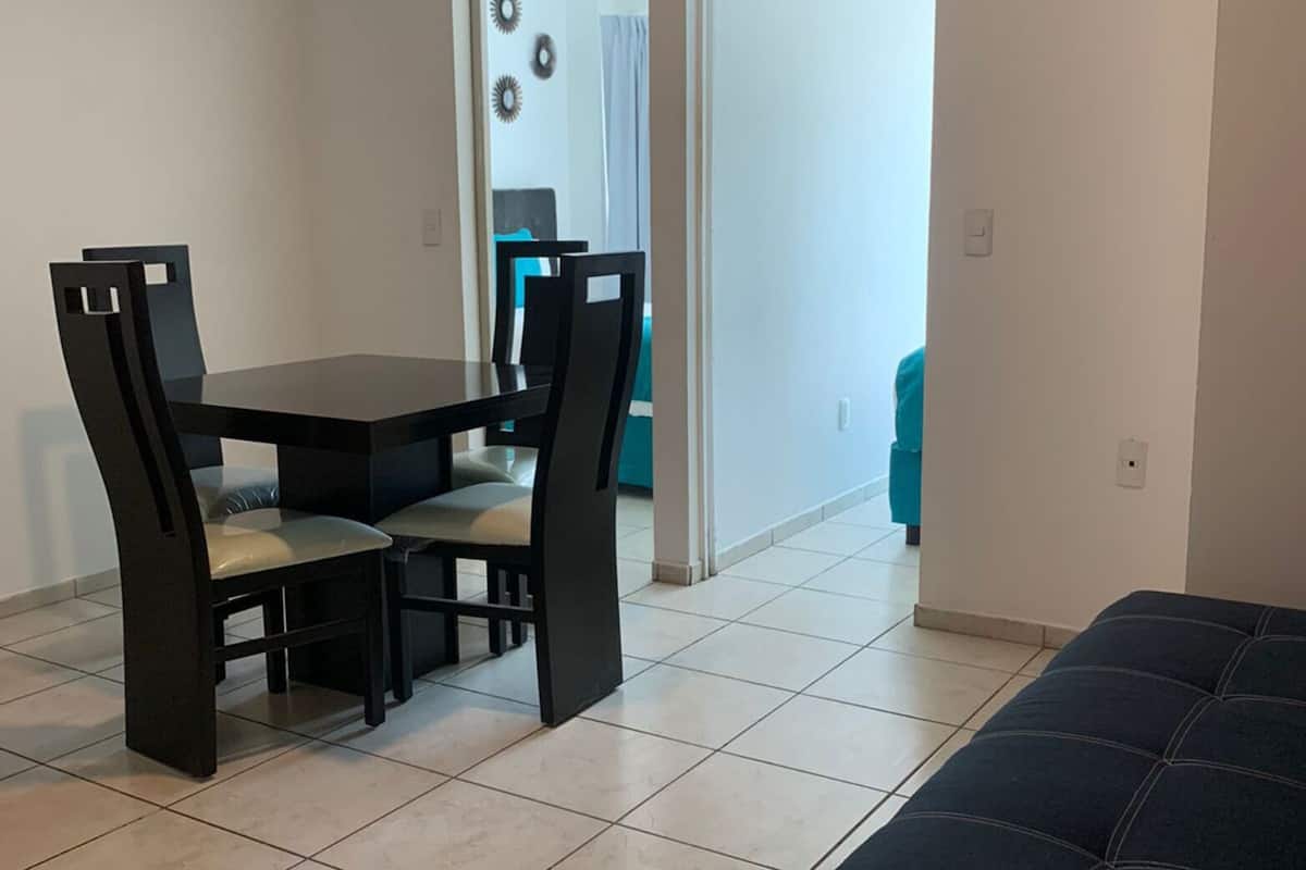 Un coin repas avec une table et des chaises, un téléviseur fixé au mur et une vue sur une autre pièce où se trouve une chaise bleue.
