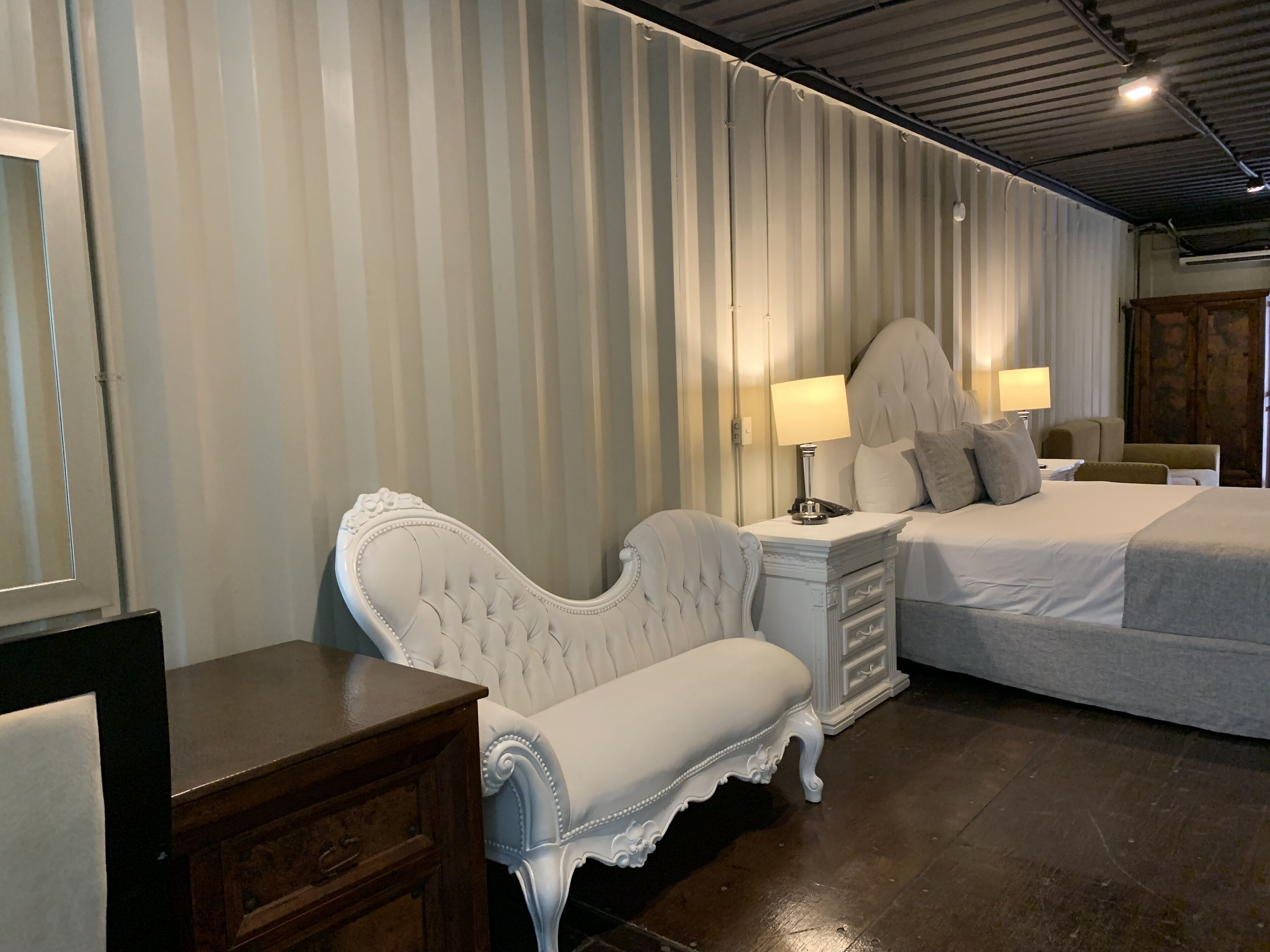 A hotel room with a large bed, a white upholstered bench, a nightstand with a lamp, and a wooden dresser.