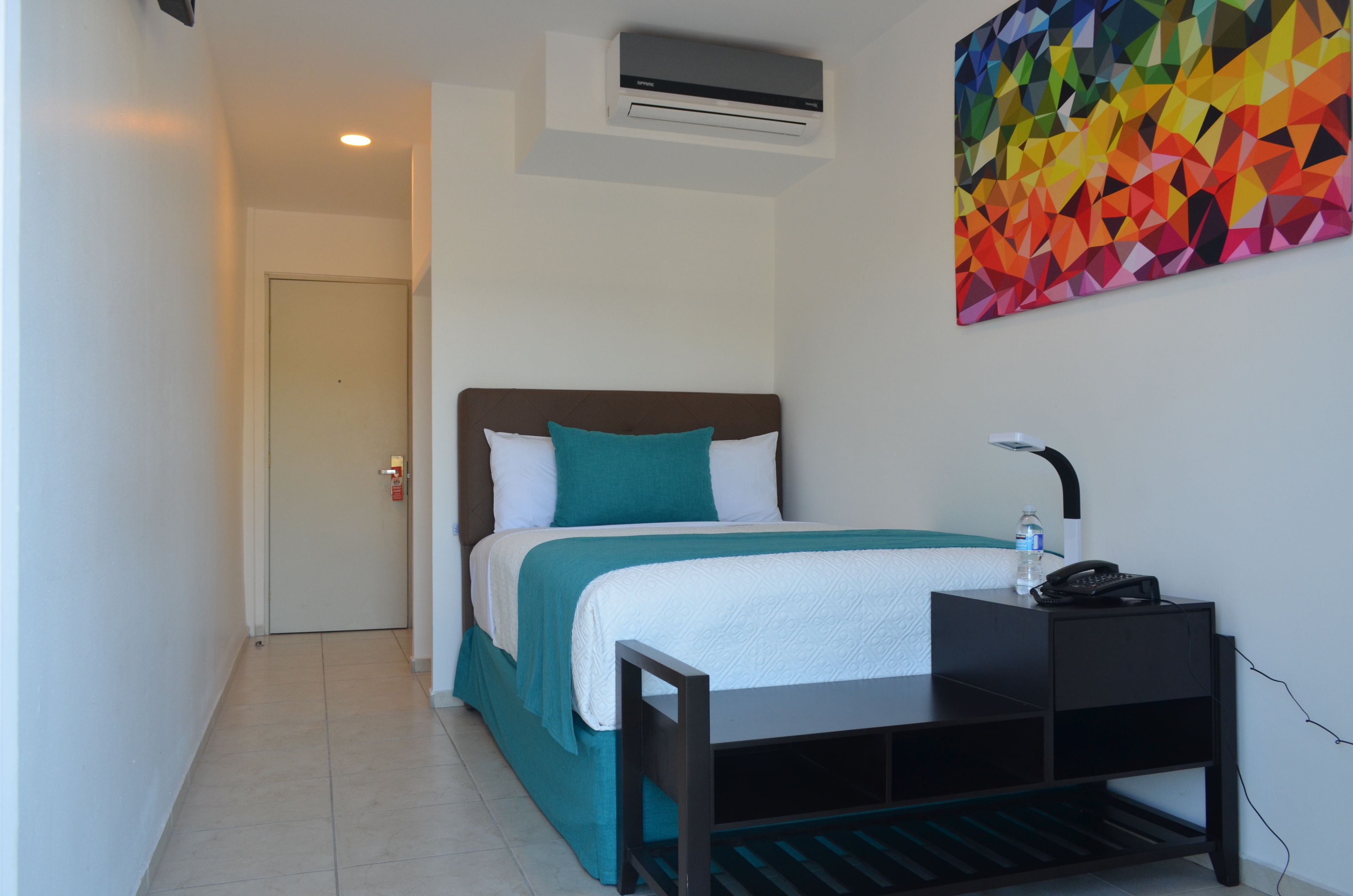 Habitación de hotel con cama, escritorio con teléfono, aire acondicionado y un cuadro de arte geométrico y colorido.