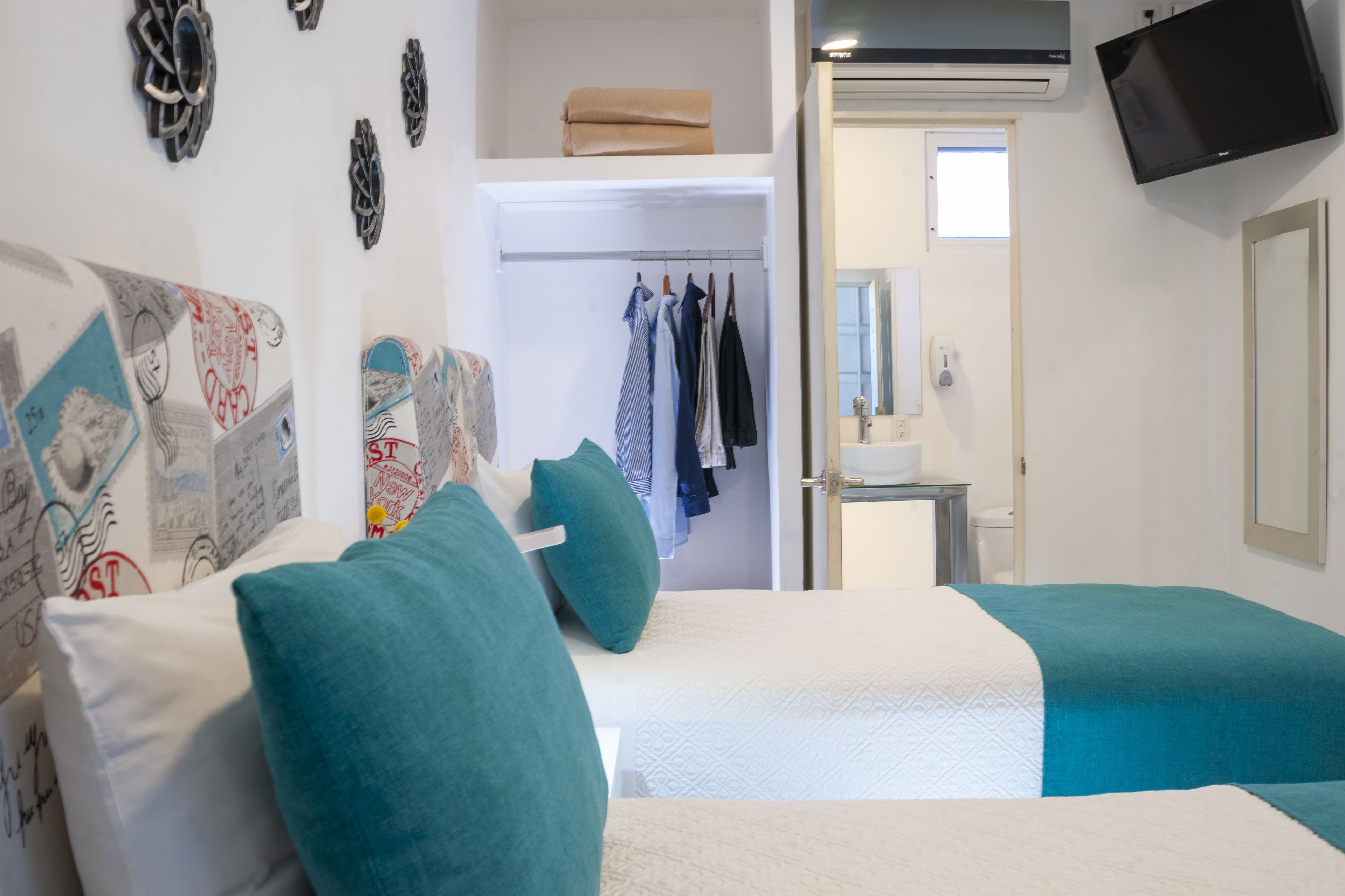 A hotel room with two beds, teal and white bedding, a wardrobe with hanging clothes, a small desk with a mirror, and a TV mounted on the wall.