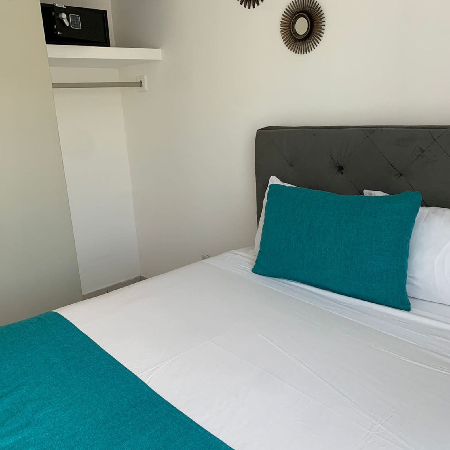 Una cama bien hecha con un cojín verde azulado y ropa de cama blanca, un cabecero tapizado gris y un pequeño estante encima de la cama con un objeto negro.