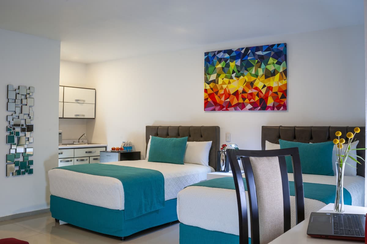 Une chambre d’hôtel moderne avec deux lits, un bureau, un ordinateur portable et une peinture abstraite aux couleurs vives accrochée au mur.