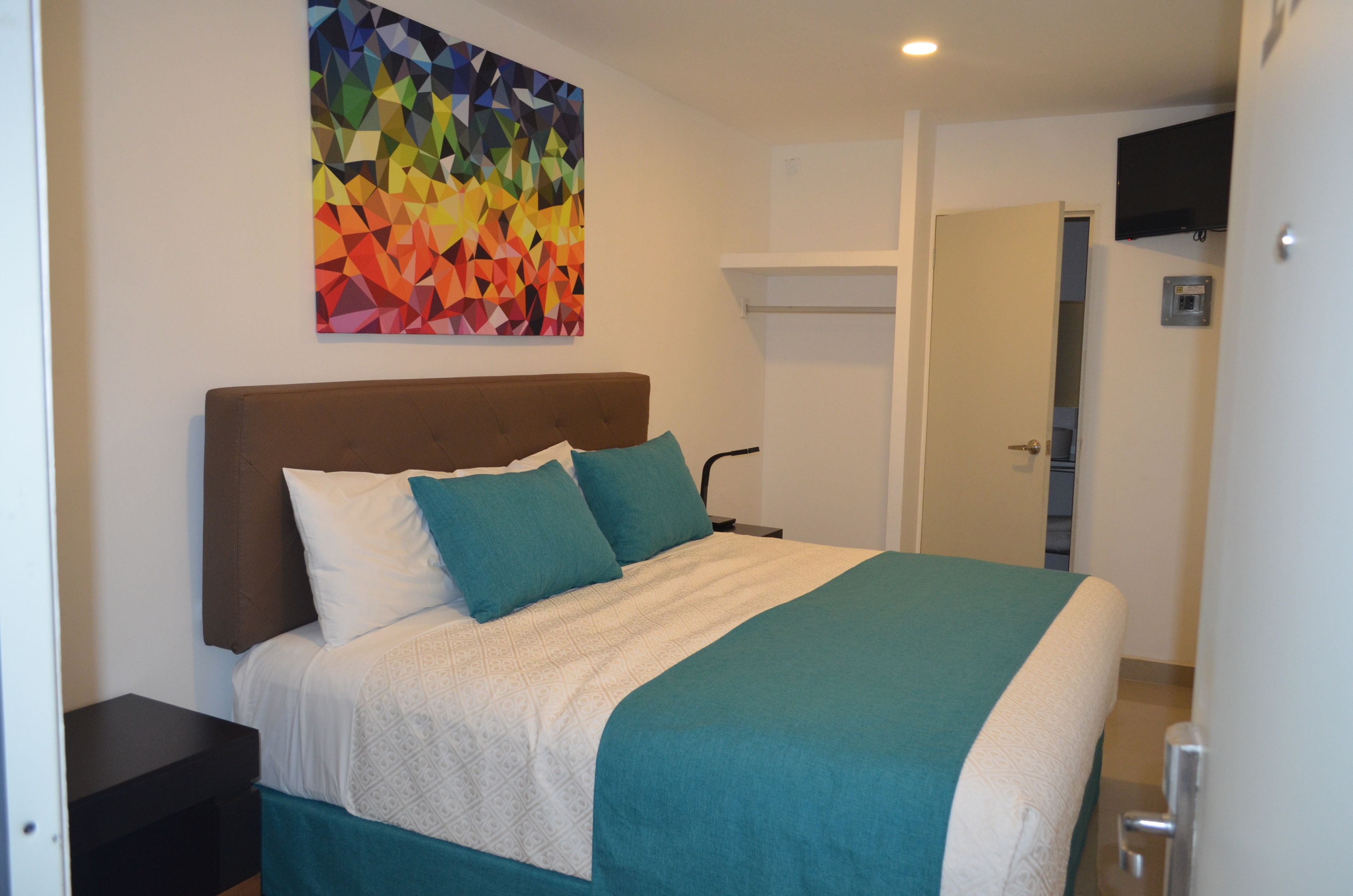 A neatly made bed with a patterned bedspread, a colorful abstract painting on the wall, and a television mounted above the bed.
