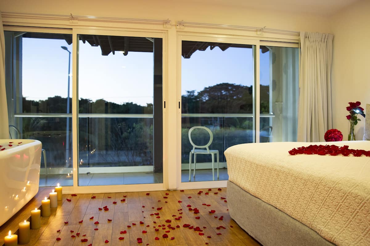 Une chambre avec une grande porte-fenêtre coulissante donnant sur un balcon, un lit parsemé de pétales de roses et une baignoire blanche.