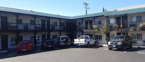 A motel with a central courtyard parking area, multiple balconies with potted plants, and a clear blue sky.