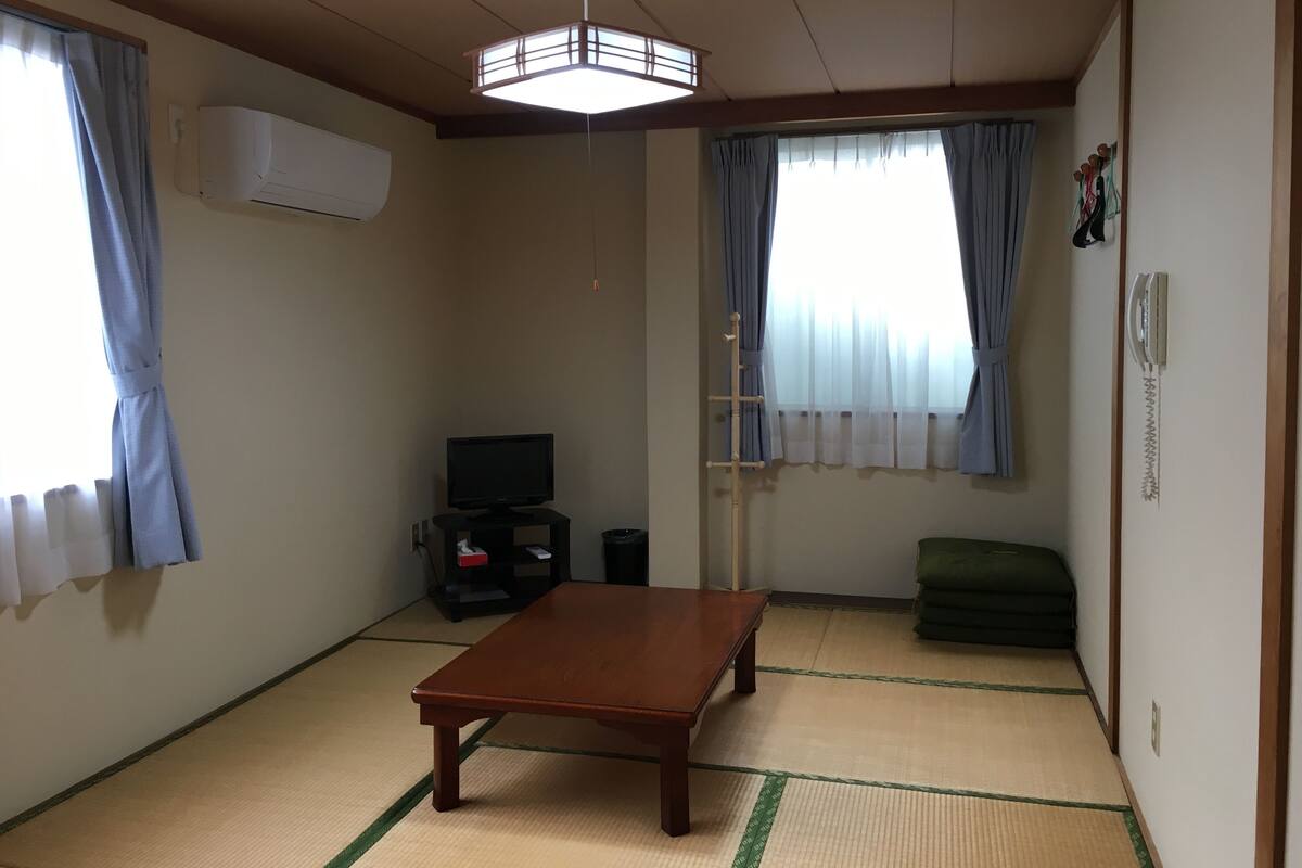 Ein Zimmer mit Tatami-Böden, einem Holztisch, einem Fernseher und einem Futon.