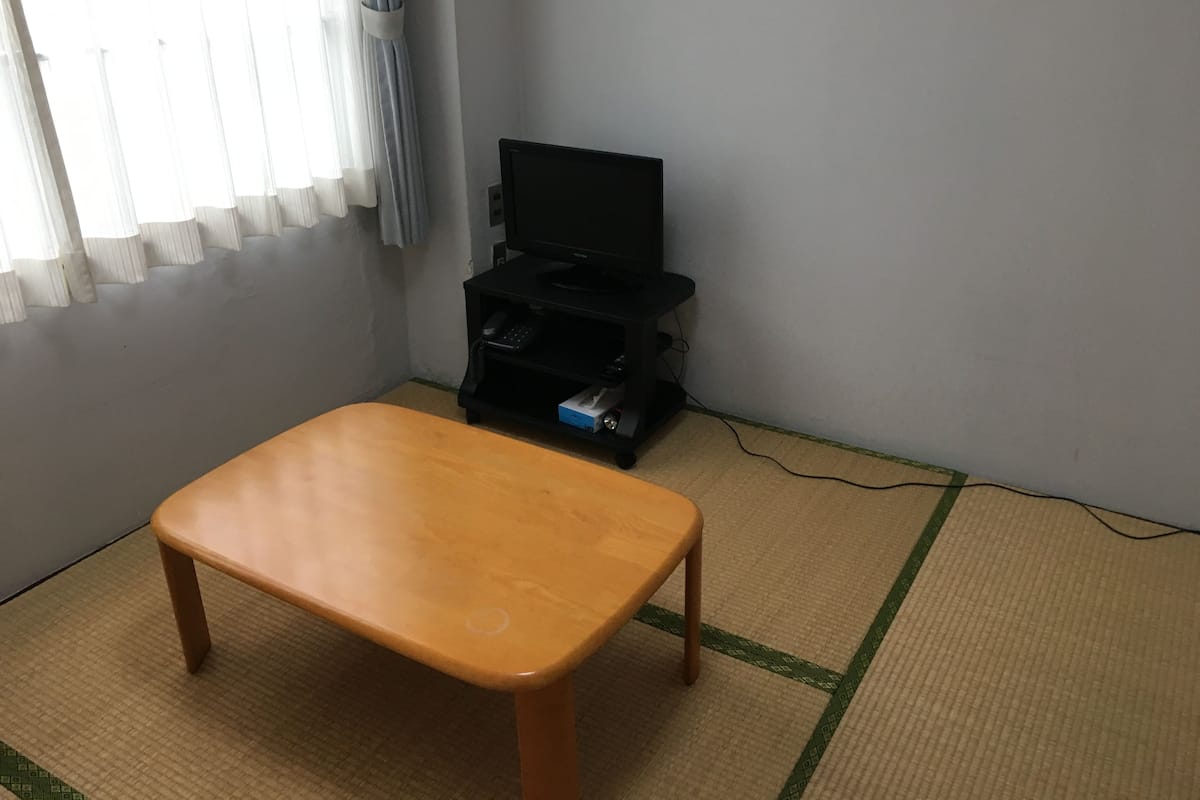 Ein Zimmer mit einem Holztisch, einer schwarzen Unterhaltungsecke mit Fernseher und einem Fenster mit Vorhängen.