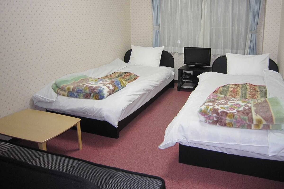 Ein Hotelzimmer mit zwei Einzelbetten, einem kleinen Tisch, einem Fernseher und einem Fenster mit Vorhängen.