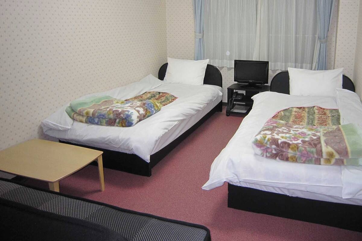 Ein Hotelzimmer mit zwei Einzelbetten, einem kleinen Tisch, einem Fernseher und einem Schreibtisch.