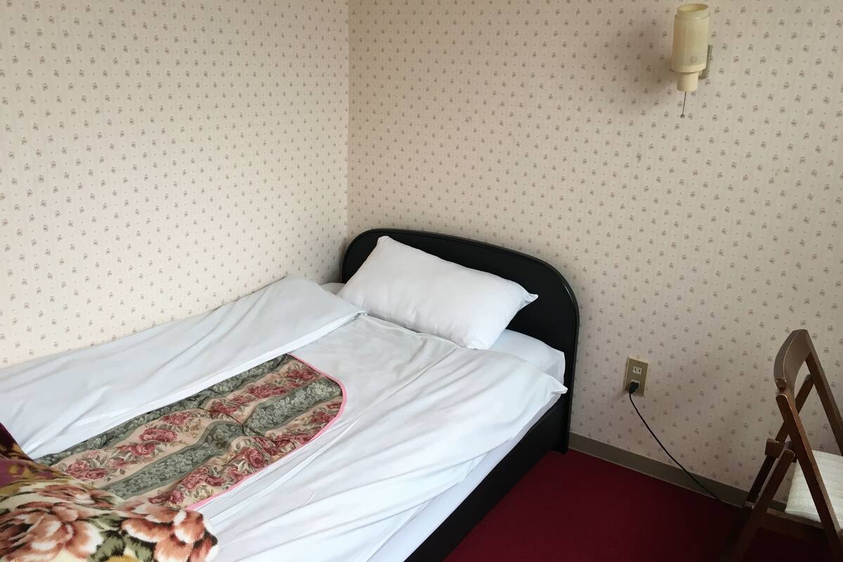 Ein Einzelbett mit weißer Bettwäsche, einer floralen Zudecke, einem schwarzen Kopfteil und einer an der Wand befestigten Leuchte.