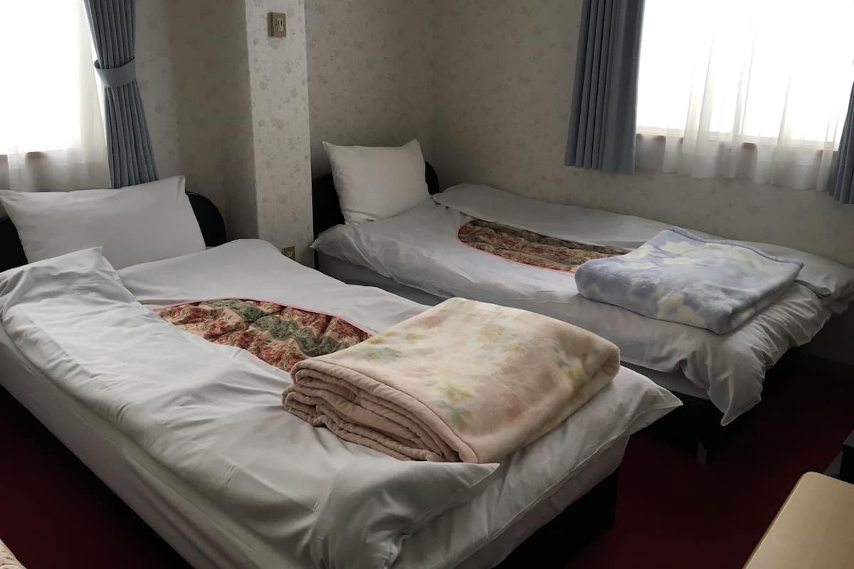 Ein Hotelzimmer mit zwei Einzelbetten, jeweils mit weißer Matratze und einer zudeckenden Decke mit Blumenmuster. Im Vordergrund steht ein kleiner Tisch, und das Zimmer hat ein schlichtes, zweckmäßiges Design mit schlichten Wänden und einem Fenster mit Vorhängen.