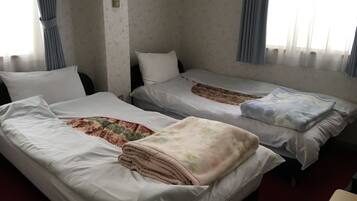 A hotel room with two single beds, each with a white mattress and a floral-patterned duvet. There is a small table in the foreground, and the room has a simple, functional design with plain walls and a window with curtains.