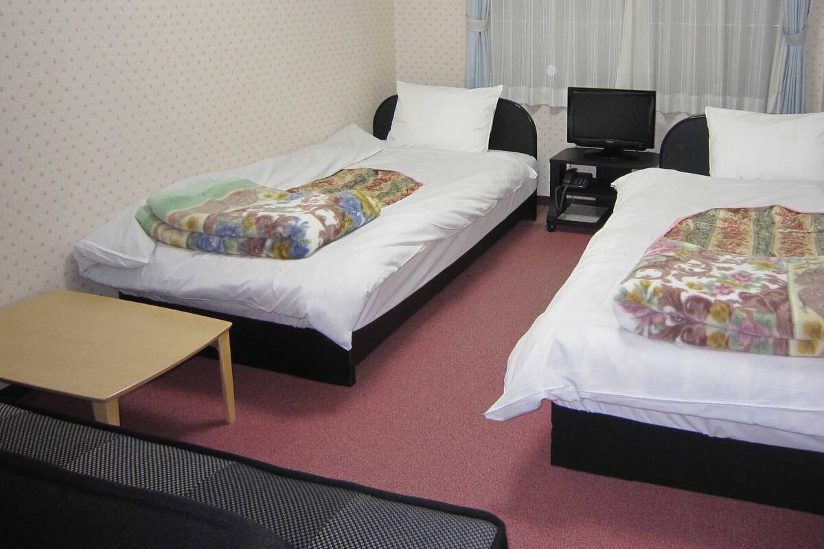 Ein Hotelzimmer mit zwei Einzelbetten, einem kleinen Tisch, einem Fernseher und einem Schreibtisch.