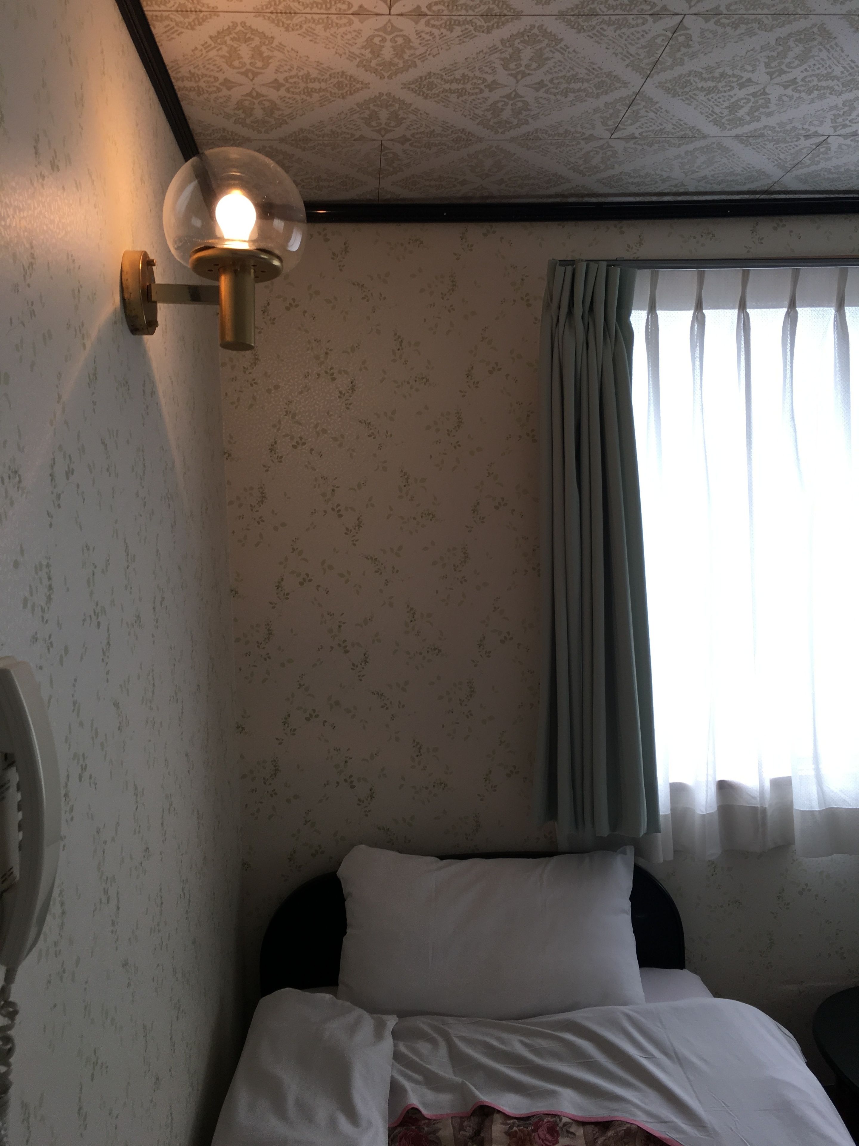 A hotel room with a bed, a wall-mounted lamp, a window with curtains, and a patterned wallpaper.