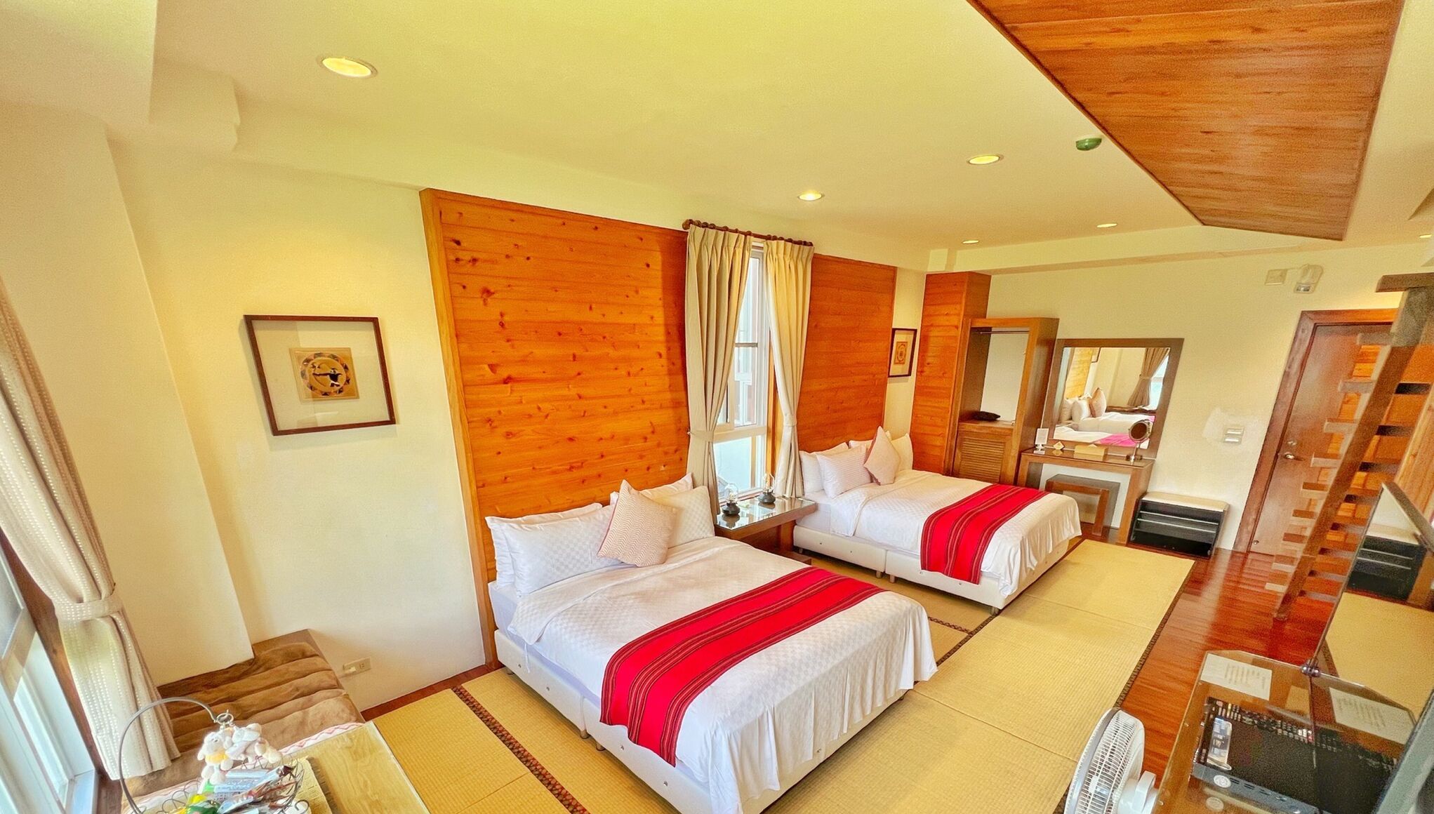 Une chambre d’hôtel avec deux lits, des panneaux de bois au mur, un grand miroir et un ventilateur.