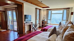 Habitación de hotel con piso de madera, televisor sobre un mueble de madera, escritorio con computadora, cama con ropa blanca y roja, y ventanas amplias con vista a la montaña.