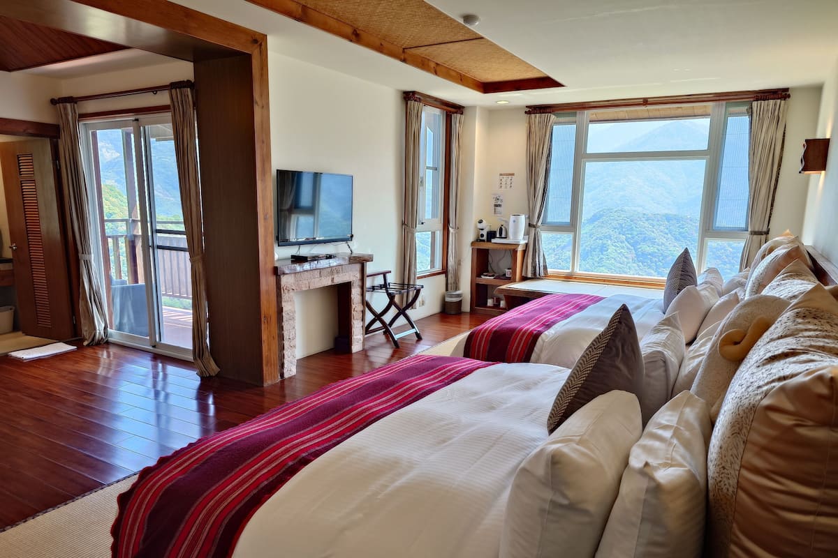 A hotel room with a wooden floor, a TV on a wooden stand, a desk with a computer, a bed with white and red bedding, and large windows with a mountain view.