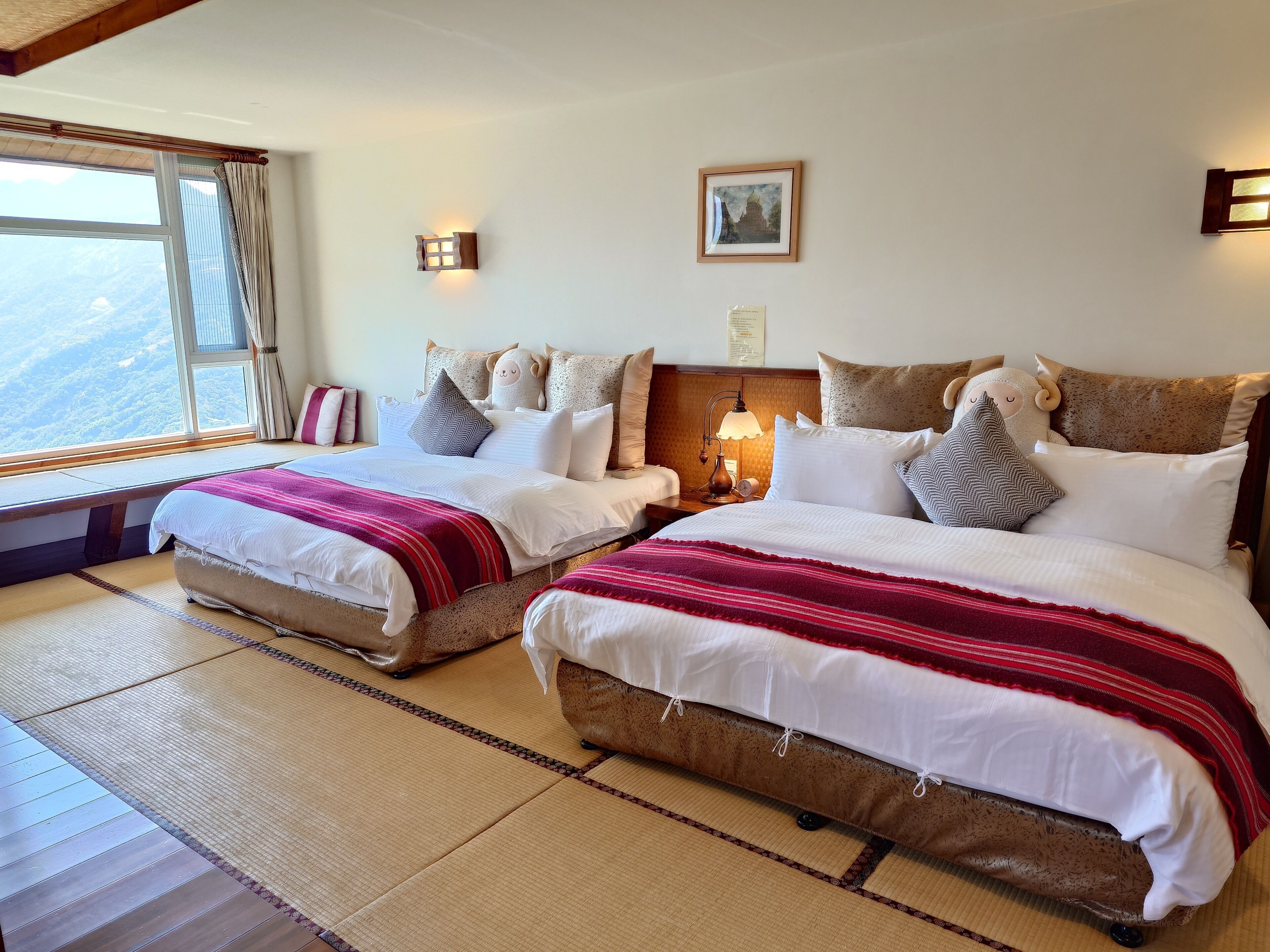 A hotel room with two beds, a large window with a view, a bedside lamp, and a framed picture on the wall.
