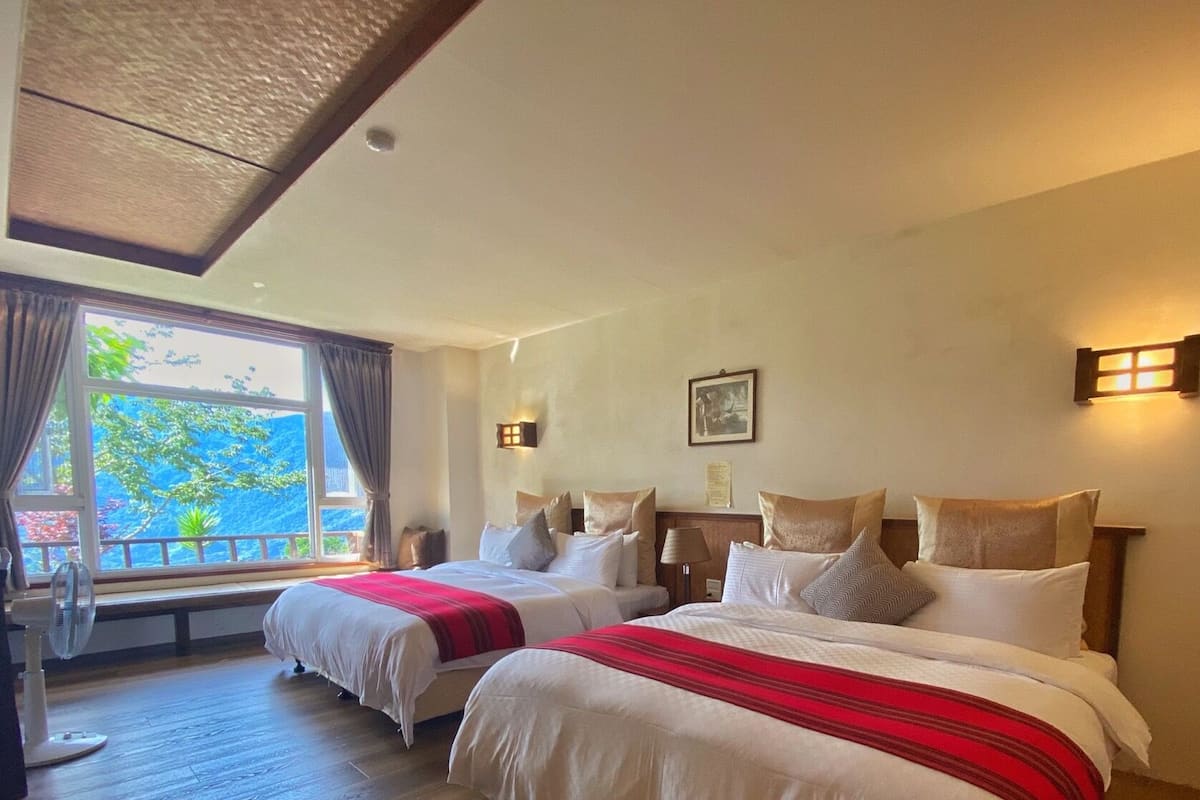 A hotel room with two beds, a large window with curtains, a ceiling fan, and a wall-mounted light fixture.