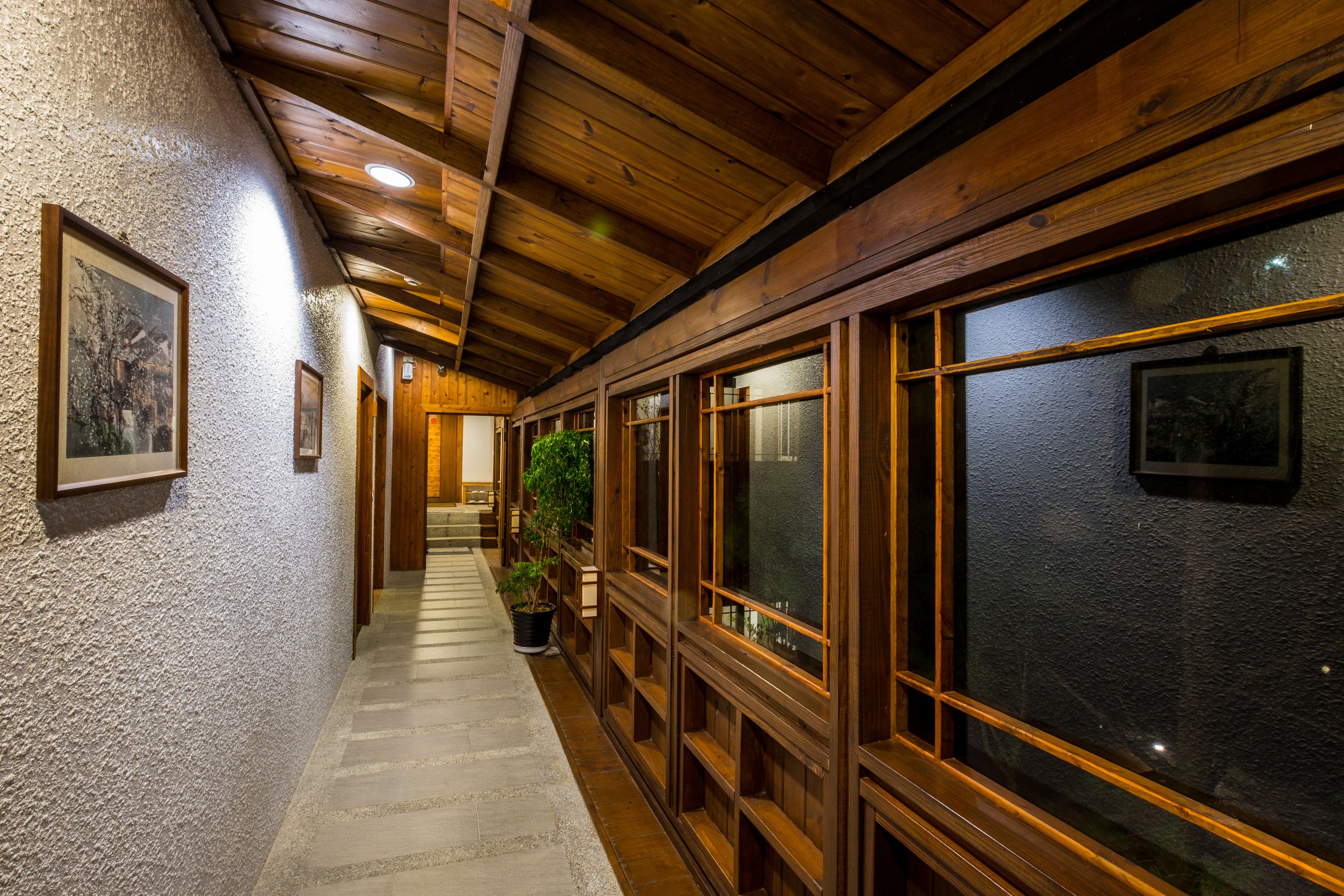 A hallway with wooden beams and a tiled floor, leading to a room with a large window and a potted plant.