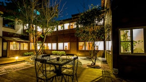 Una terraza de madera con una mesa y sillas, rodeada de árboles y un edificio con ventanas iluminadas.