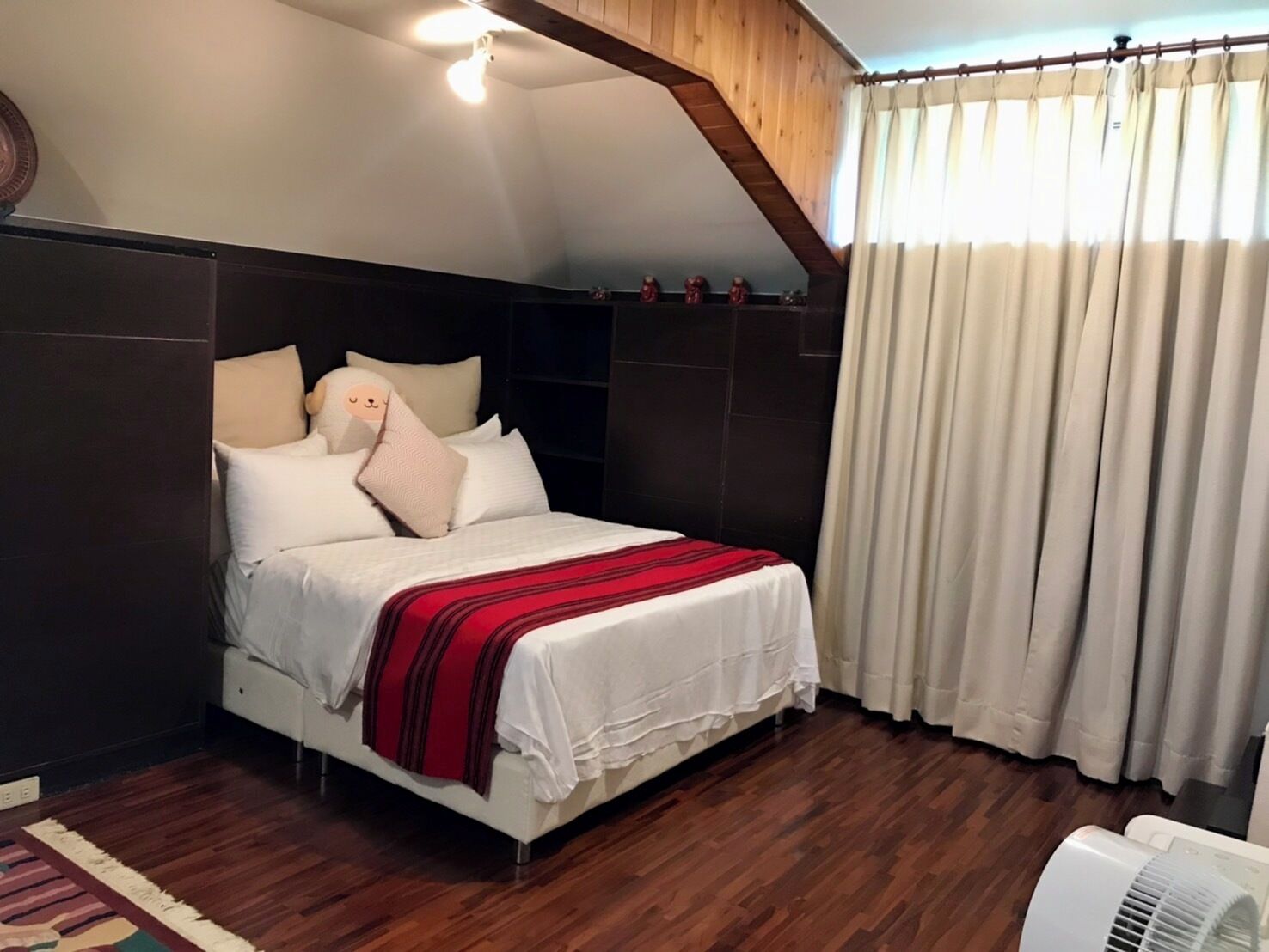 A bedroom with a sloped ceiling, a bed with white bedding and a red blanket, wooden paneling on the walls, and a ceiling fan.