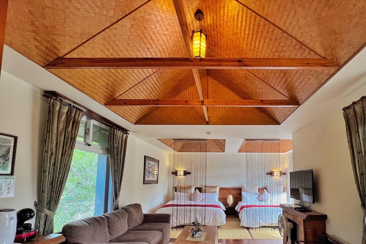 A hotel room with a wooden ceiling, two beds with white linens, a sofa, a TV, and large windows with curtains.