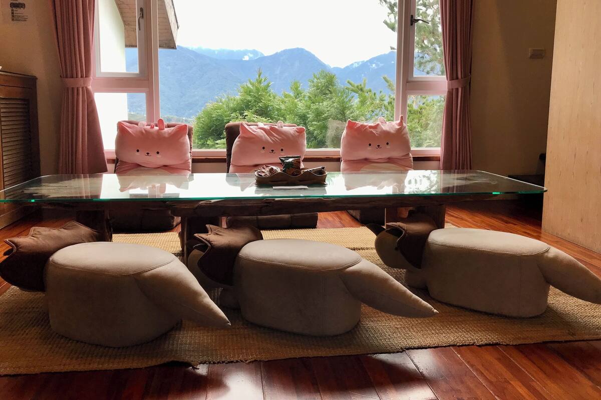 A room with a glass table, three cat-shaped cushions, and a view of mountains through large windows.