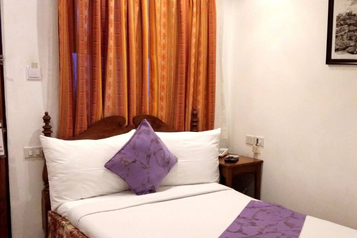 A hotel room with a wooden door, a bed with white and purple bedding, a nightstand with a phone, and a framed picture on the wall.