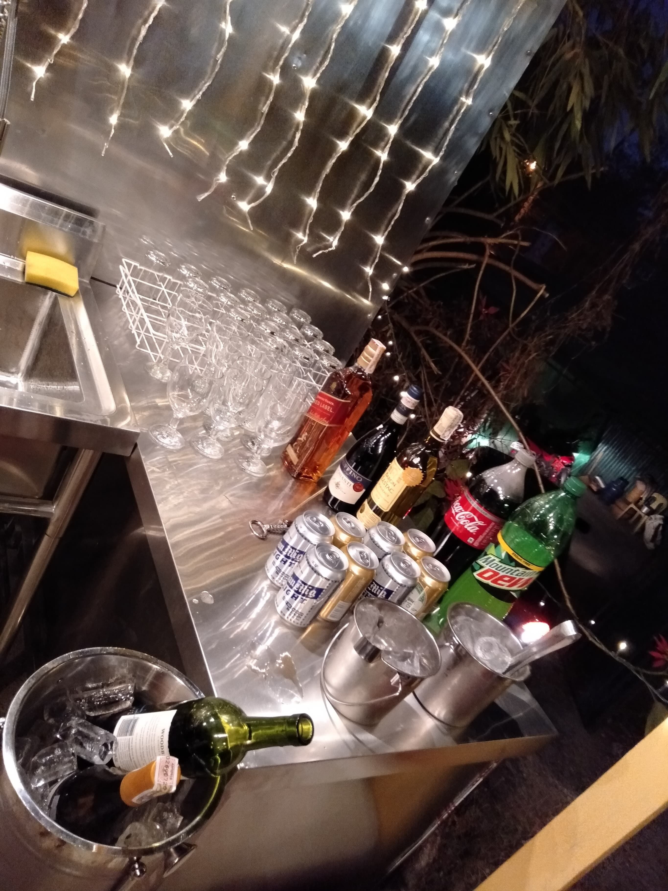 A bar area with a stainless steel counter, glasses, bottles, and cans.