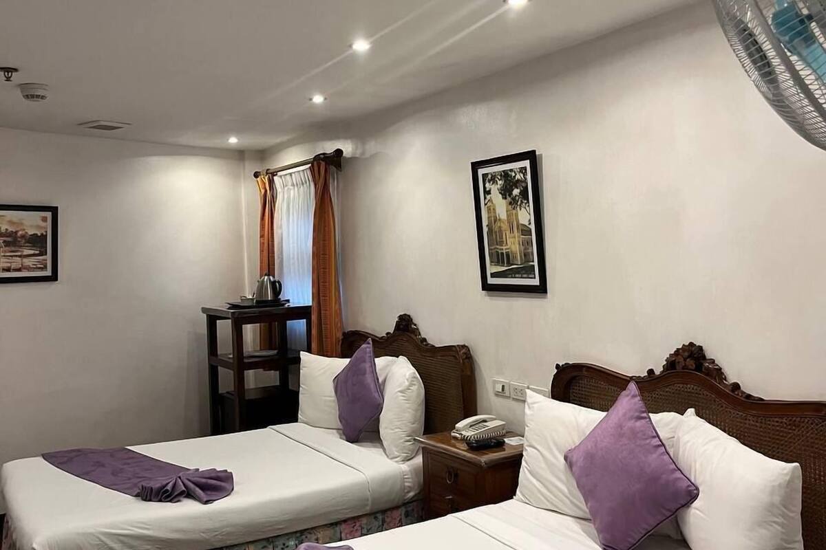A hotel room with two beds, a desk, a chair, a nightstand, a fan, and two framed pictures on the wall.