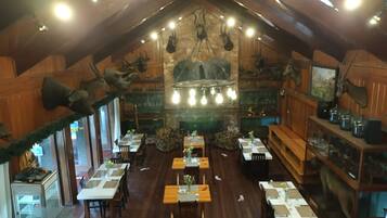 Un restaurante con paredes de madera, una chimenea de piedra y cuernos de ciervo colgados. Las mesas están dispuestas con manteles blancos y centros de mesa.