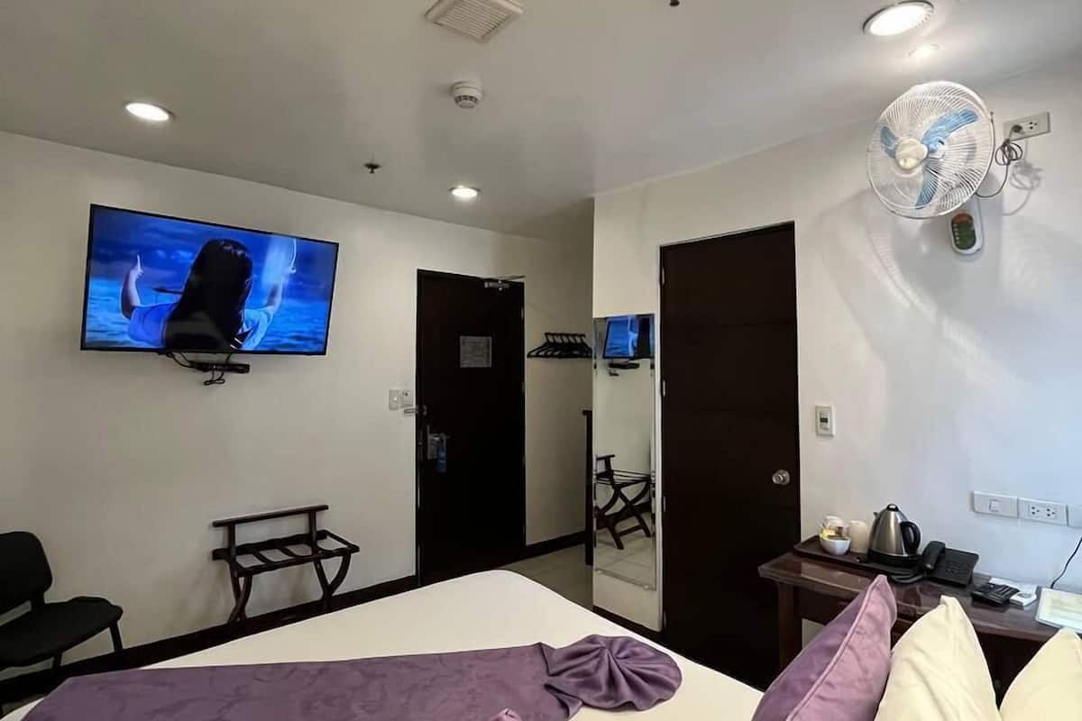 A hotel room with a bed, a TV mounted on the wall, a desk with a kettle, and a fan.