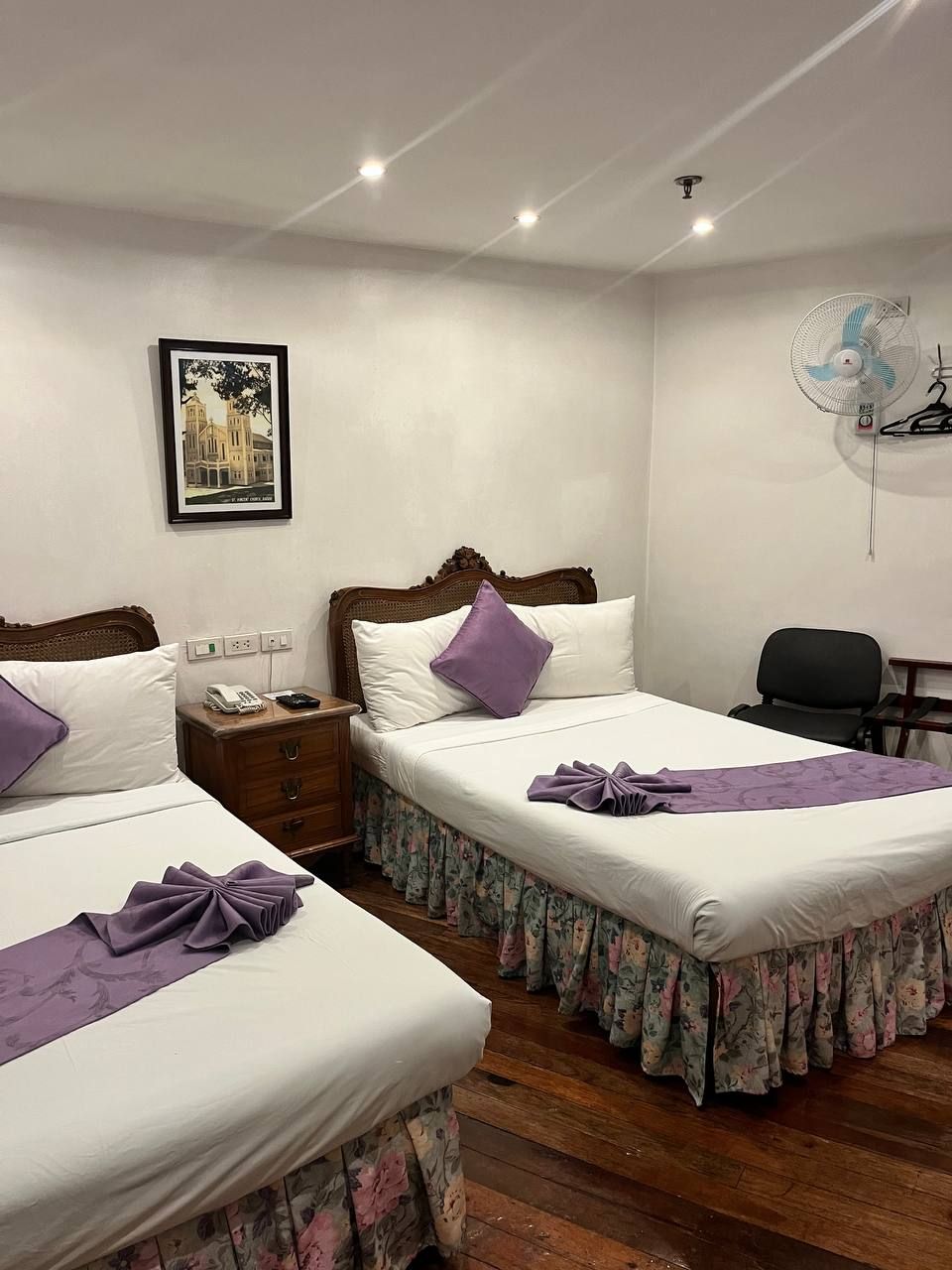 Een hotelkamer met twee bedden, een houten hoofdbord, een nachtkastje met een telefoon, een lijst met een foto aan de muur en een bureau met een stoel.